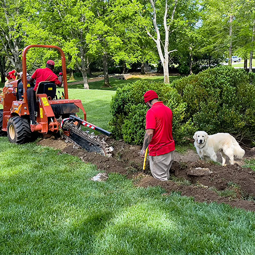 Two technicians use a shovel and a trenching machine to dig in a customer's yard while the homeowner's pet watches.
