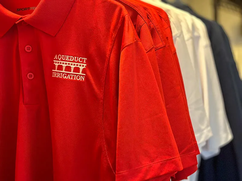 A row of hanging shirts in red white and blue with the Aqueduct Irrigation logo embroidered on to them.